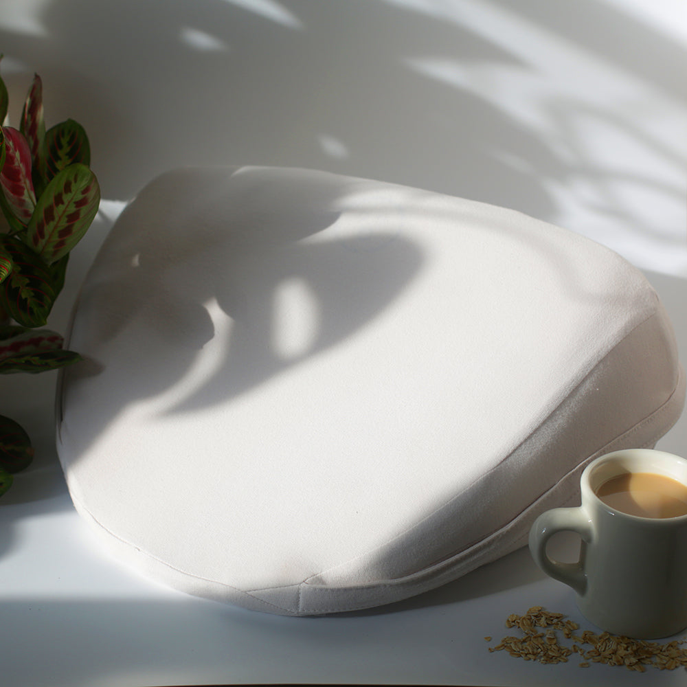 Dame Pillo Sex Position Pillow Oat on a surface with a cup of coffee and greenery in the background.