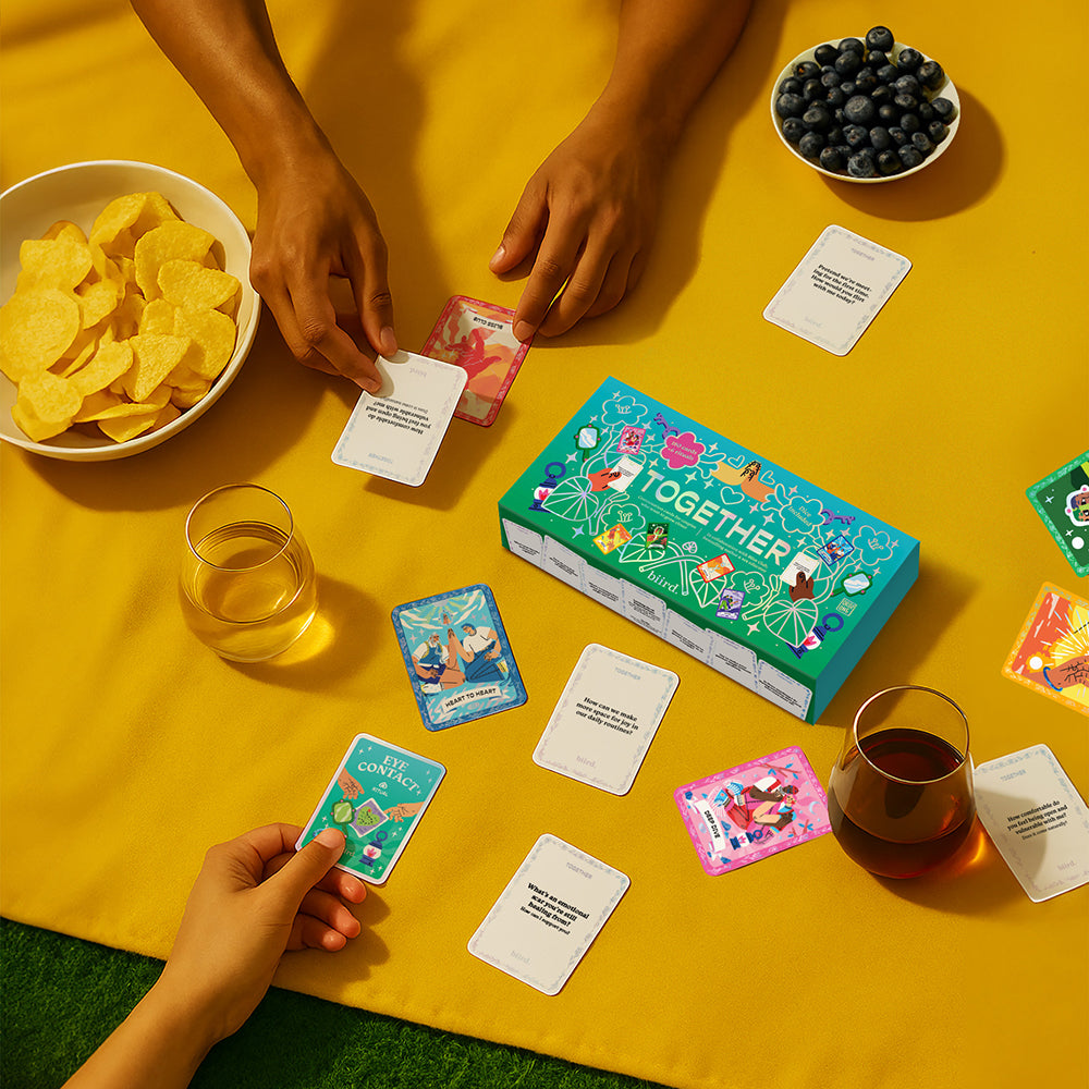 Biird X Jouissance Club Together Couples Card Game displayed with food and drinks on a yellow blanket.