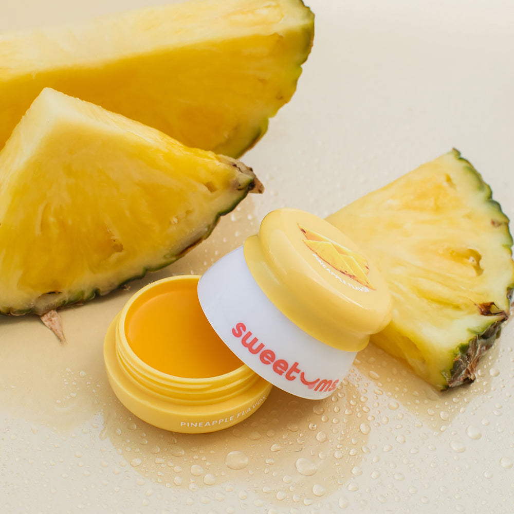 Sweetums Pineapple Flavored Lip Balm opened next to fresh pineapple slices on a reflective surface.