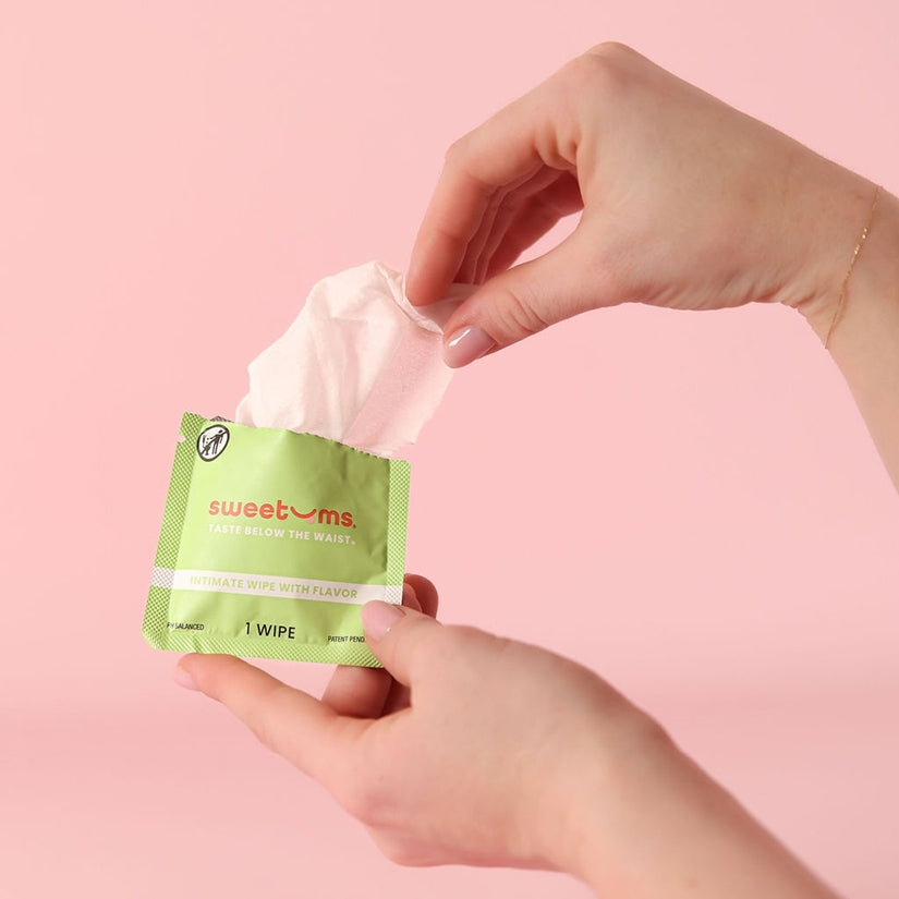 Sweetums Flavored Intimate Wipes Watermelon being pulled from a green packet on a pink background.